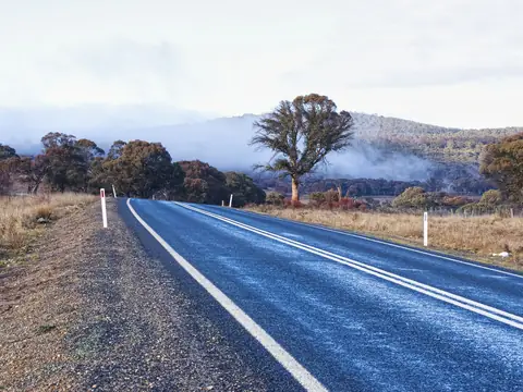 A sealed country road with white guide posts curving past dry grass and gum trees, with mist lying over the wooded hills behind