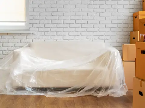 A sofa covered in clear plastic sheeting against a white brick wall, beside a stack of cardboard moving boxes and a tape dispenser
