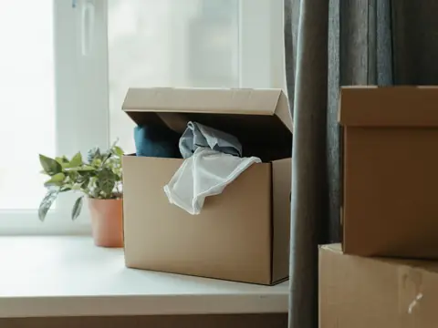A cardboard box of folded clothes on a white window sill beside a potted plant, with more boxes stacked by a grey curtain