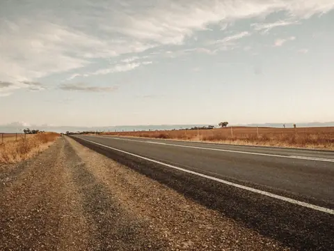 A straight sealed country road running into the distance past dry golden paddocks under a pale sky