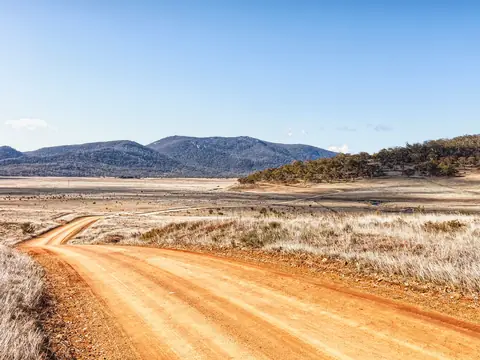 A red dirt road curving through pale dry grass towards wooded hills under a clear sky