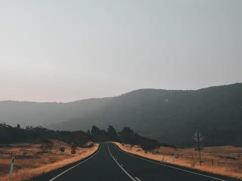 An empty two-lane road curving between dry paddocks towards wooded hills in a soft haze