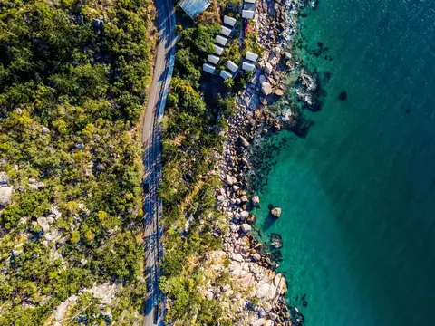 Aerial view of a coast road running through green bush beside granite boulders and clear blue water